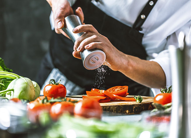 Koch würzt Tomaten vom Foodservice Ein Koch in Schürze würzt geschnittene Tomaten auf einem Holzbrett, frische Tomaten und Gemüse vom Foodservice im Vordergrund.